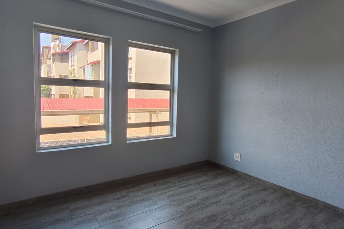 A bright, empty bedroom featuring modern grey wood-look laminate flooring, neutral painted walls, and dual windows providing natural light.