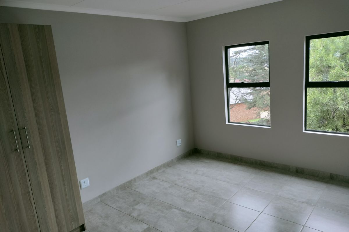 A bright, neutral-toned bedroom featuring large windows, tiled flooring, and a built-in wooden wardrobe.