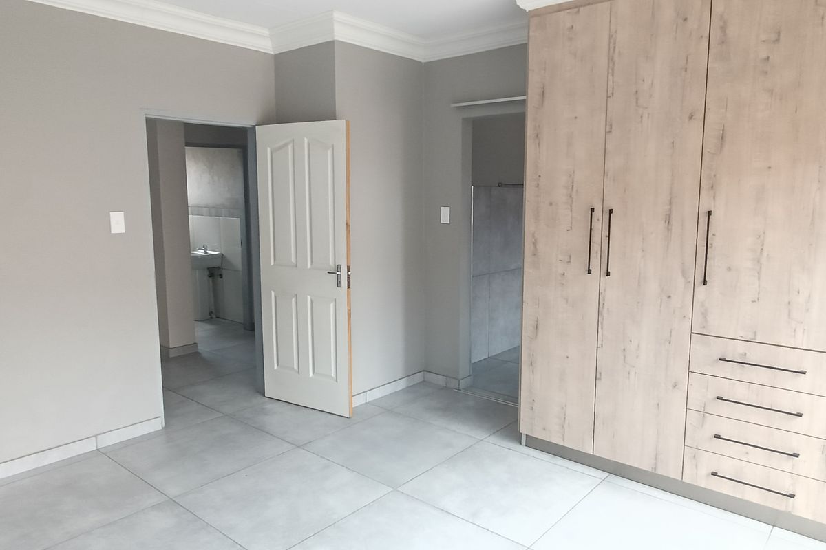 A bright bedroom featuring modern light-wood built-in cupboards, neutral grey wall paint, and large format floor tiles.