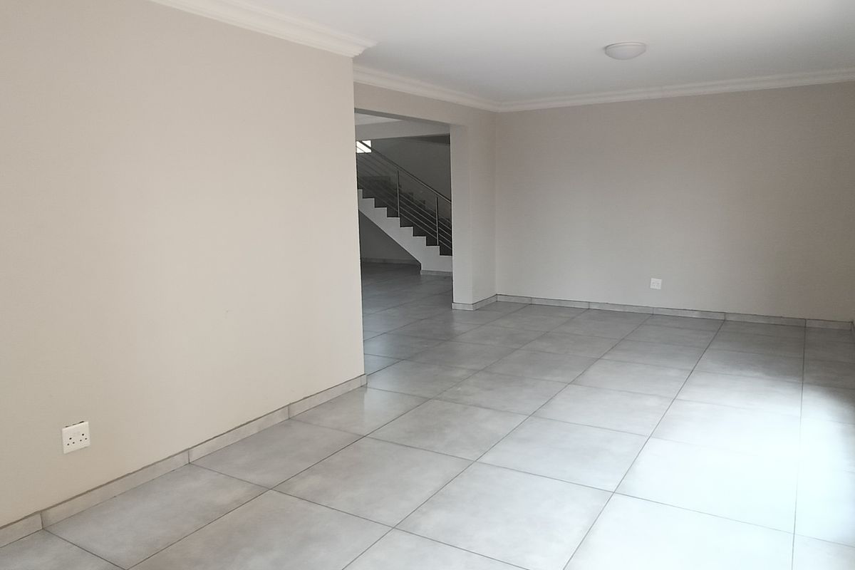 Spacious, open-plan living area featuring neutral tiled flooring, coved ceilings, and large windows with security bars, leading to an internal staircase.
