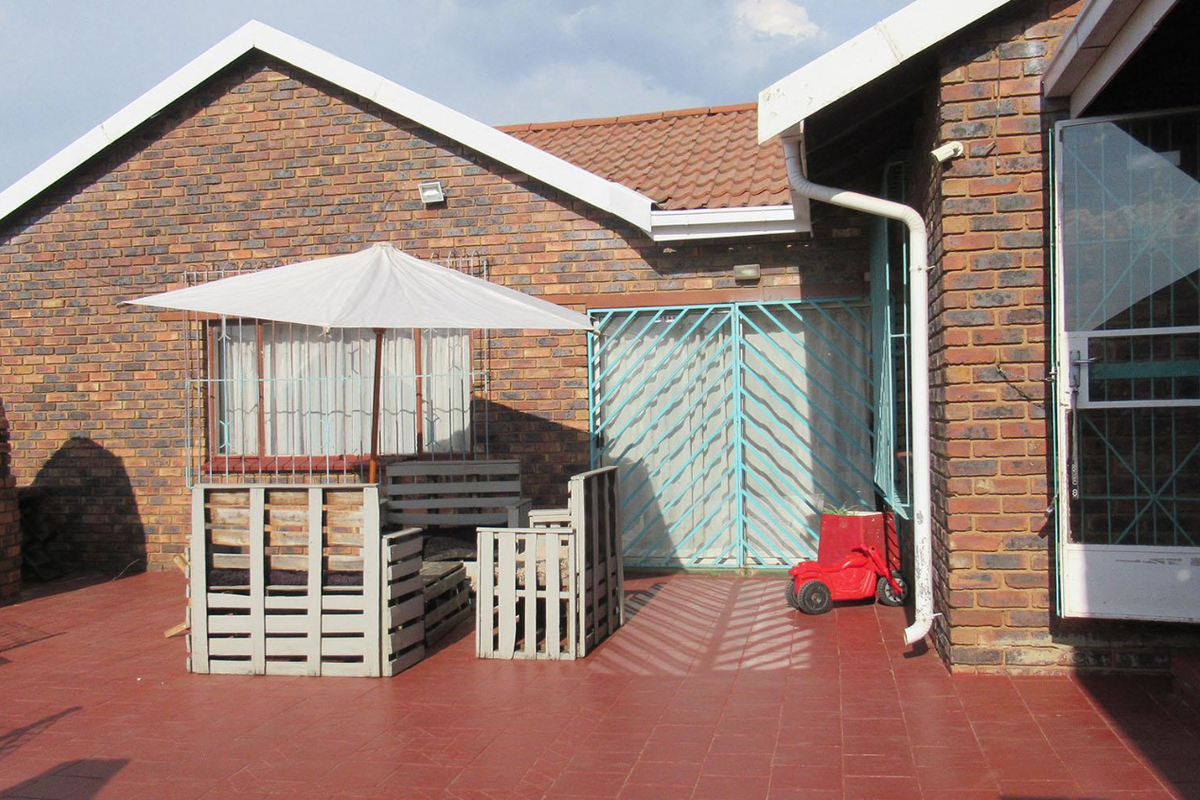 A rear patio area featuring red tiled flooring, face brick exterior walls, and blue metal security gates providing access to the home.