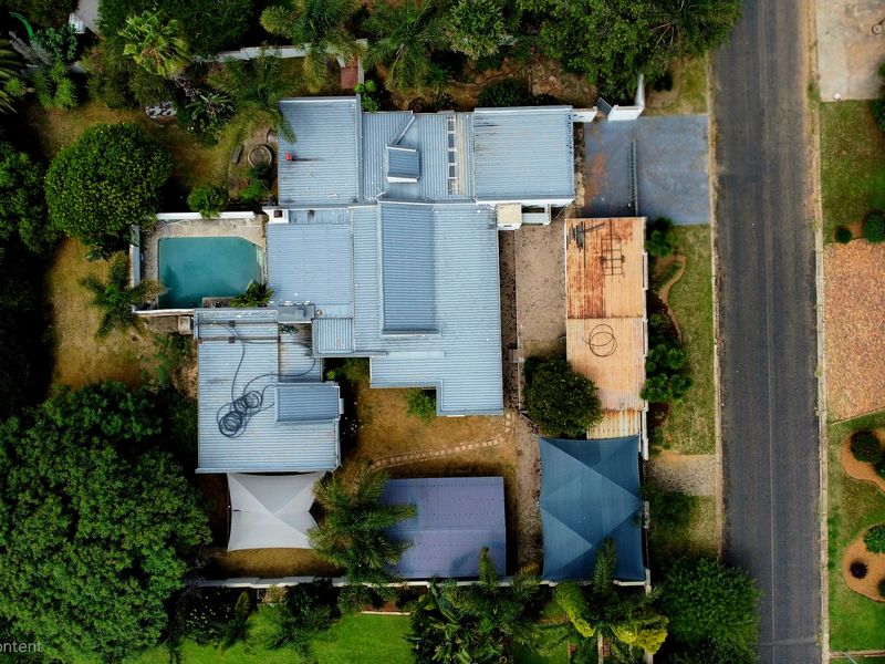 Aerial view of a residential property featuring a metal roof, swimming pool, paved driveway, and established garden areas.