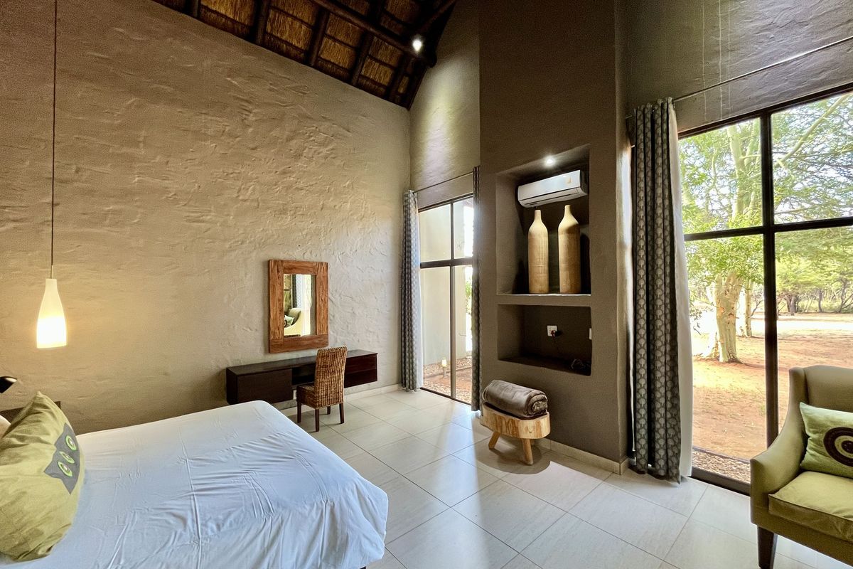 Spacious bedroom featuring high thatched ceilings, large windows with garden views, tiled flooring, and a built-in wall niche with integrated air conditioning unit.