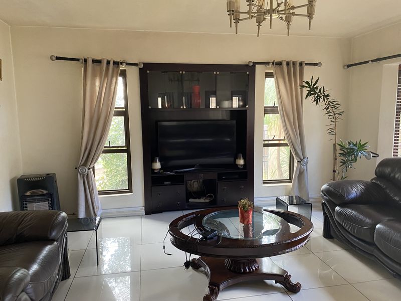 A living room featuring large white floor tiles, a central chandelier, and a dark wood built-in media cabinet flanked by two windows.