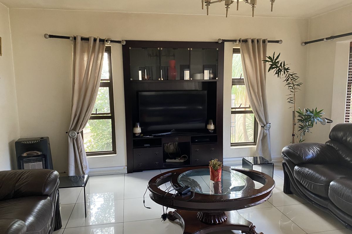 A living room featuring large white floor tiles, a central chandelier, and a dark wood built-in media cabinet flanked by two windows.