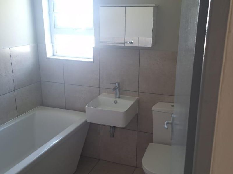 A compact, modern bathroom featuring a bathtub, wall-mounted basin, toilet, and neutral wall tiling.