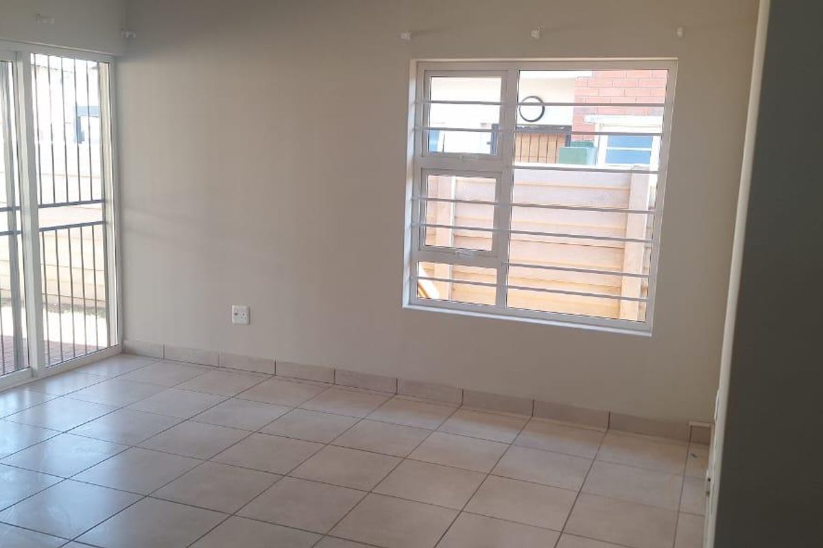 An empty living room featuring neutral tiled flooring, a sliding glass door with security bars, and a window providing natural light.