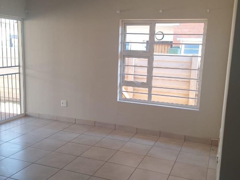 An empty living room featuring neutral tiled flooring, a sliding glass door with security bars, and a window providing natural light.