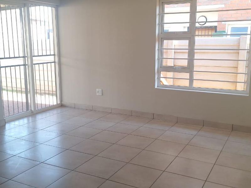 An empty living room featuring neutral tiled flooring, large windows with security bars, and a sliding glass door providing access to the exterior.