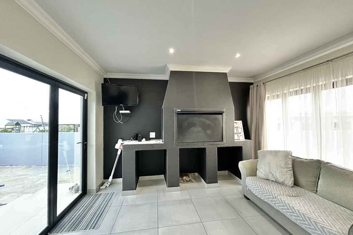 A bright living area featuring a built-in indoor braai fireplace, large glass sliding doors leading to the patio, and modern tiled flooring.