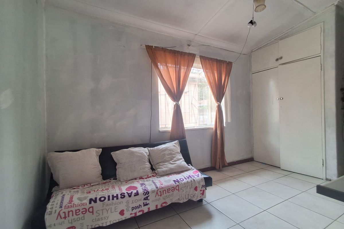 A bedroom featuring tiled flooring, built-in cupboards, and a window with security bars, offering a functional space in need of cosmetic updates.