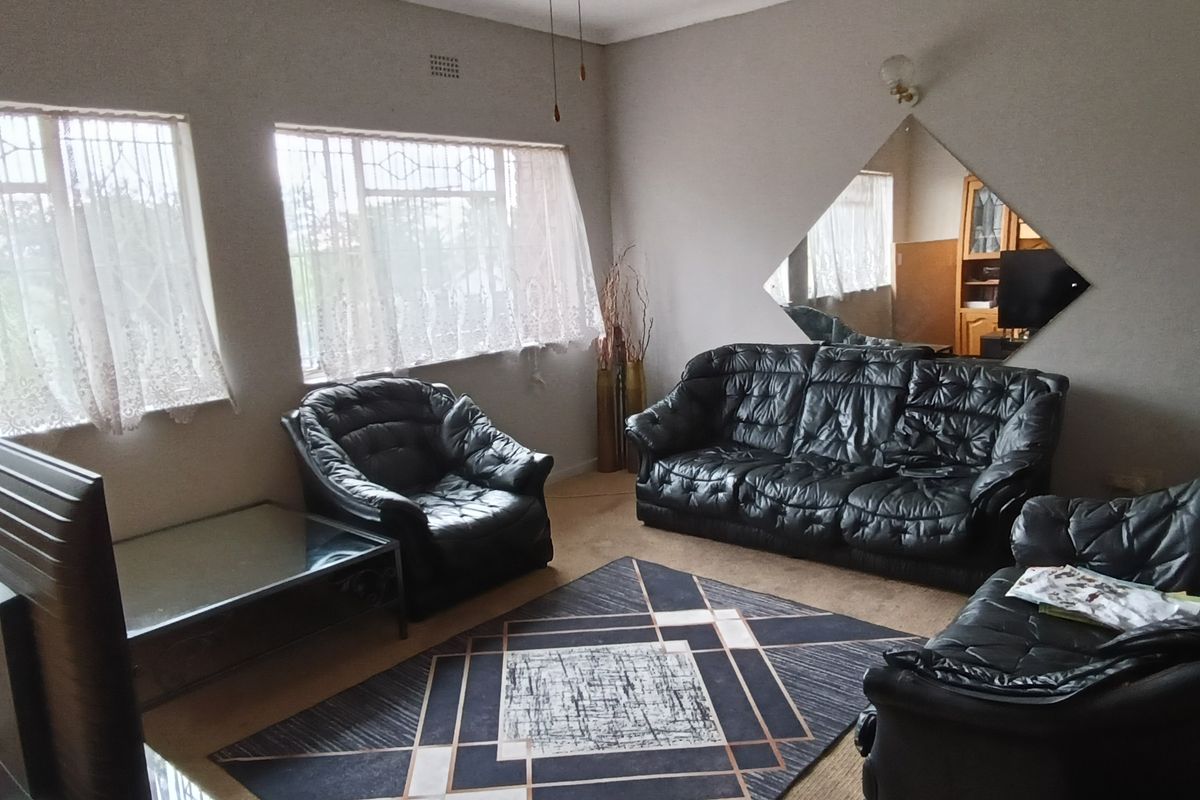 A living room featuring large windows with security bars, carpeted flooring, and a wall-mounted diamond-shaped mirror.