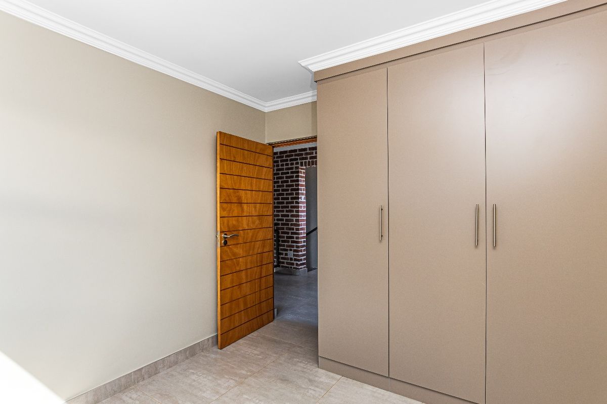 A clean bedroom interior featuring built-in floor-to-ceiling wardrobes, tiled flooring, and crown molding, with a wooden door leading to an adjacent hallway.