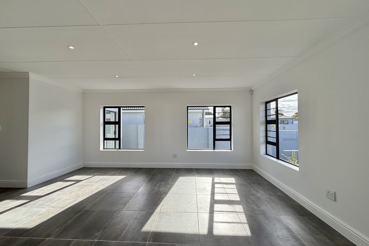 A bright, spacious, and empty living area featuring modern dark tiled flooring, crisp white walls, and large aluminium-framed windows.