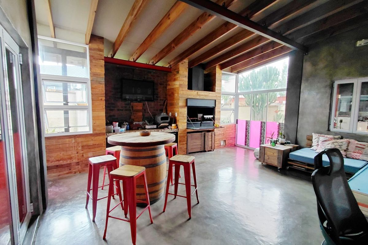 Spacious entertainment area featuring exposed timber ceiling beams, polished concrete flooring, built-in braai, and large windows providing ample natural light.
