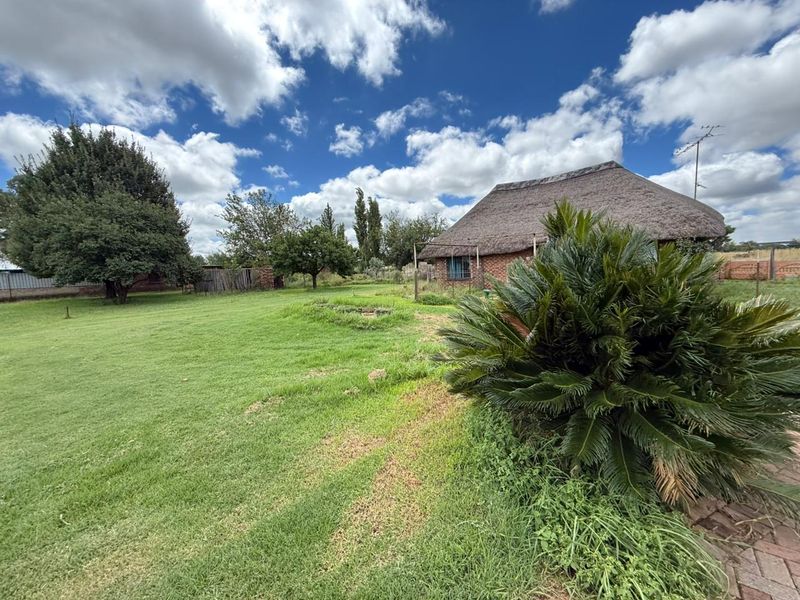A spacious garden area featuring a large lawn, mature trees, and a view of a traditional thatched-roof house exterior.