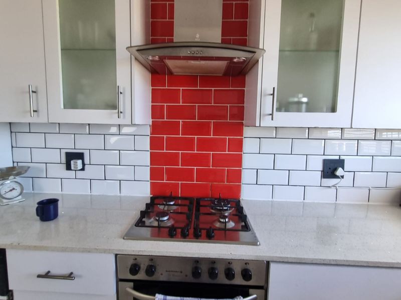 A modern kitchen featuring white cabinetry, stone countertops, a stainless steel gas hob, built-in oven, and a striking red tiled splashback.