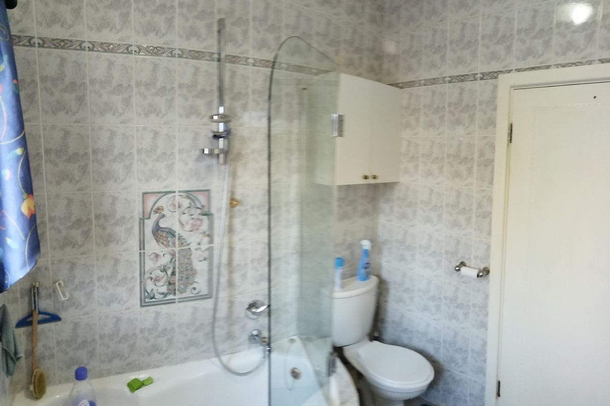 A functional bathroom featuring a bathtub with an integrated shower, glass screen, toilet, and wall-mounted storage cabinet with fully tiled walls.