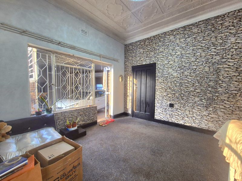 Living room featuring an ornate pressed metal ceiling, stone-effect feature wall, and security-barred windows, offering a dated interior space with potential for modernization.