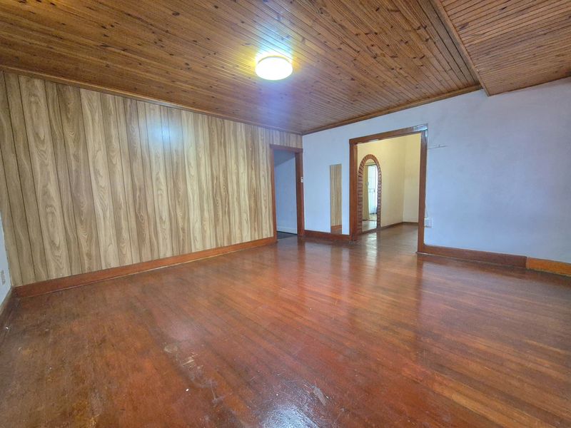 An empty living room featuring traditional timber ceiling, wood wall paneling, and hardwood flooring, leading through an archway to an adjacent space.
