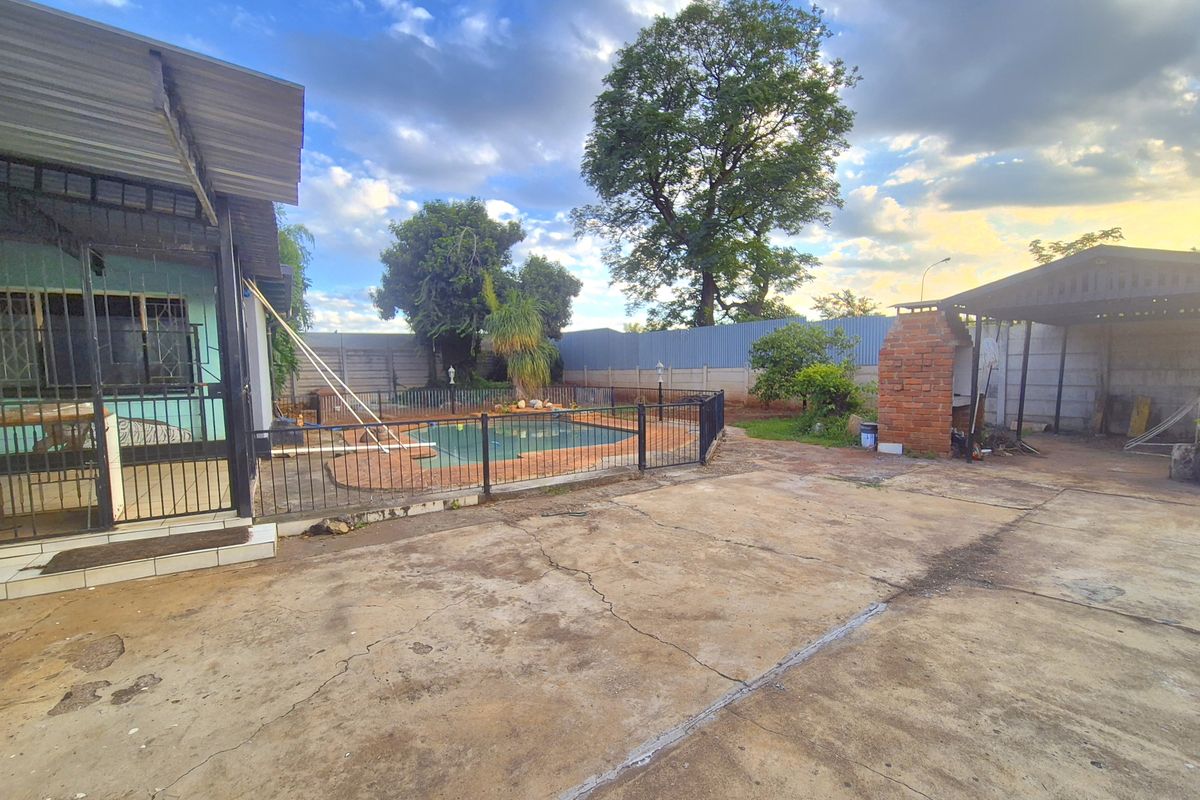 Rear exterior area featuring a swimming pool, brick braai, covered patio, and extensive concrete paving in a spacious backyard setting.