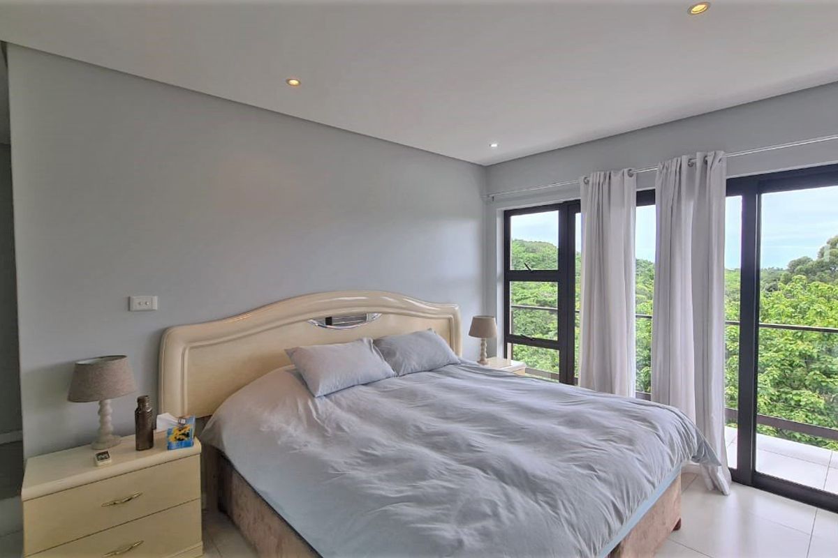 A bright bedroom featuring large sliding glass doors that open onto a balcony with scenic green views and recessed ceiling lighting.