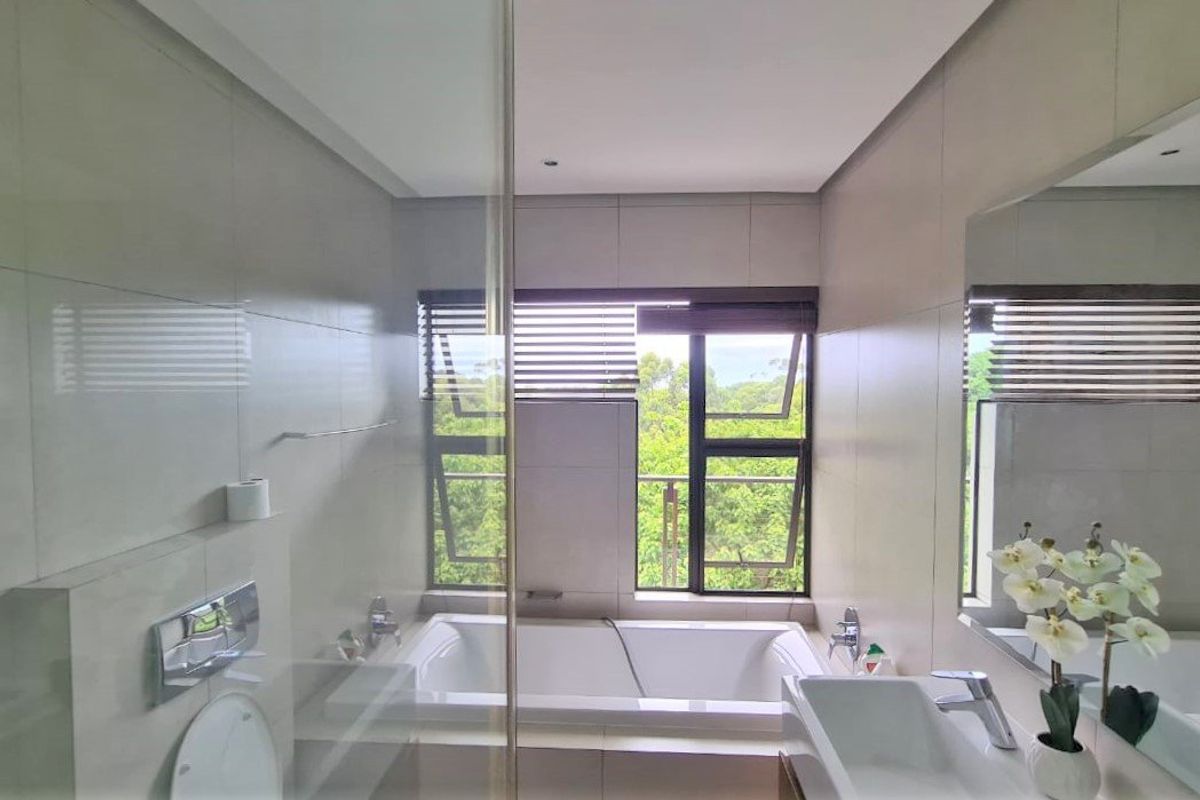 Modern bathroom featuring a built-in bathtub, wall-mounted toilet, and a sleek vanity with a large mirror, offering a pleasant view through the window.