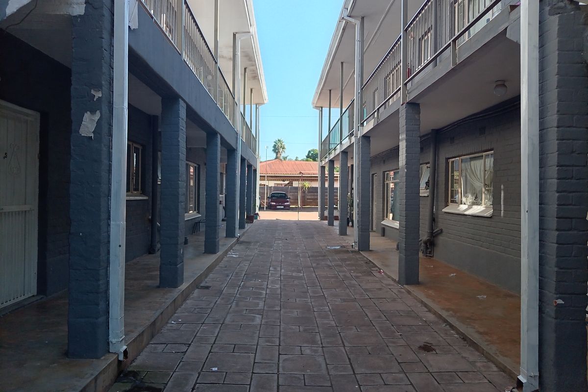 Exterior view of an apartment complex walkway featuring paved ground, covered corridors, and metal railings between two residential building blocks.
