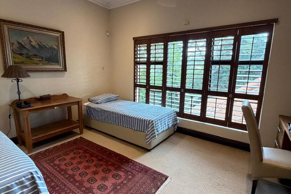 A bedroom featuring neutral carpeting, a ceiling fan, wall-mounted air conditioning unit, and large wooden shutter windows providing natural light.