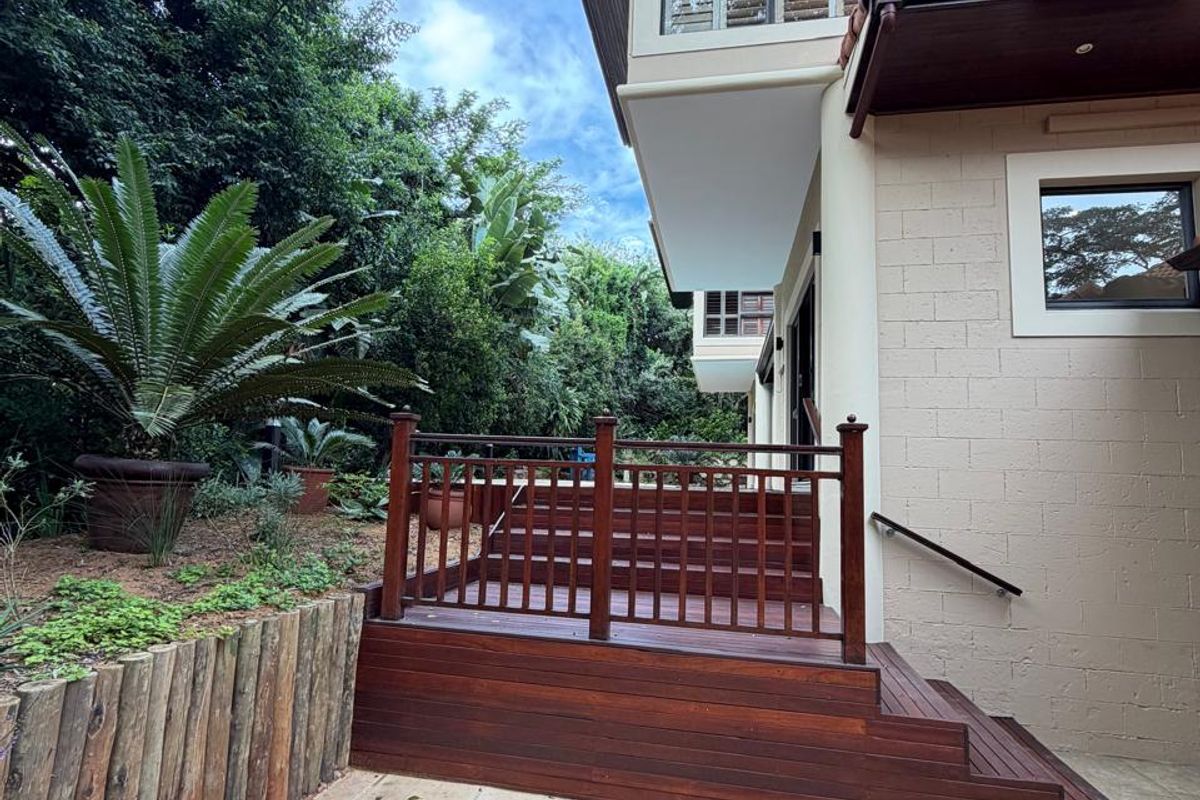 A well-maintained rear exterior featuring a timber deck with balustrades, stone paved patio area, and established garden landscaping.
