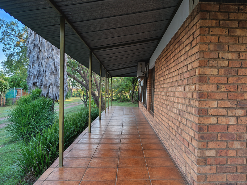 A covered outdoor patio featuring tiled flooring, exposed brick walls, and a corrugated metal roof, overlooking the surrounding garden area.
