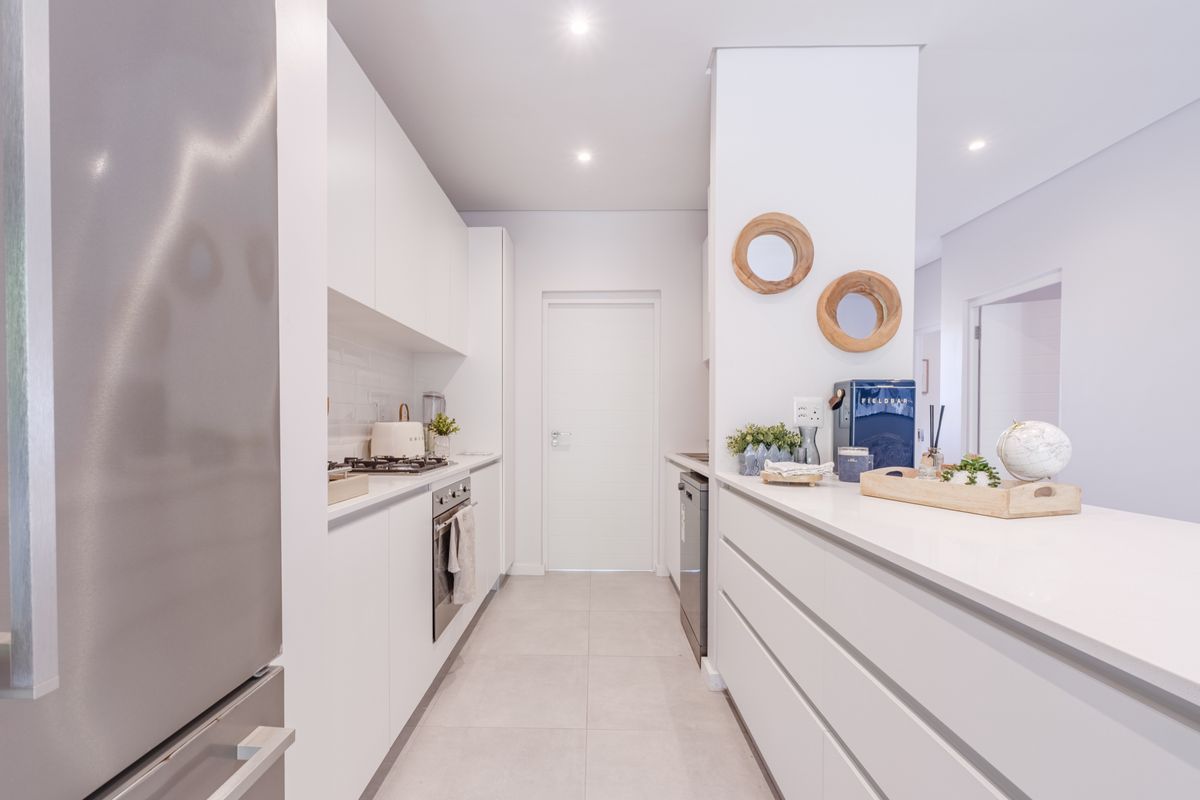 Modern galley-style kitchen featuring sleek white cabinetry, stone countertops, integrated oven, gas hob, and tiled flooring in a bright, contemporary retirement village unit.