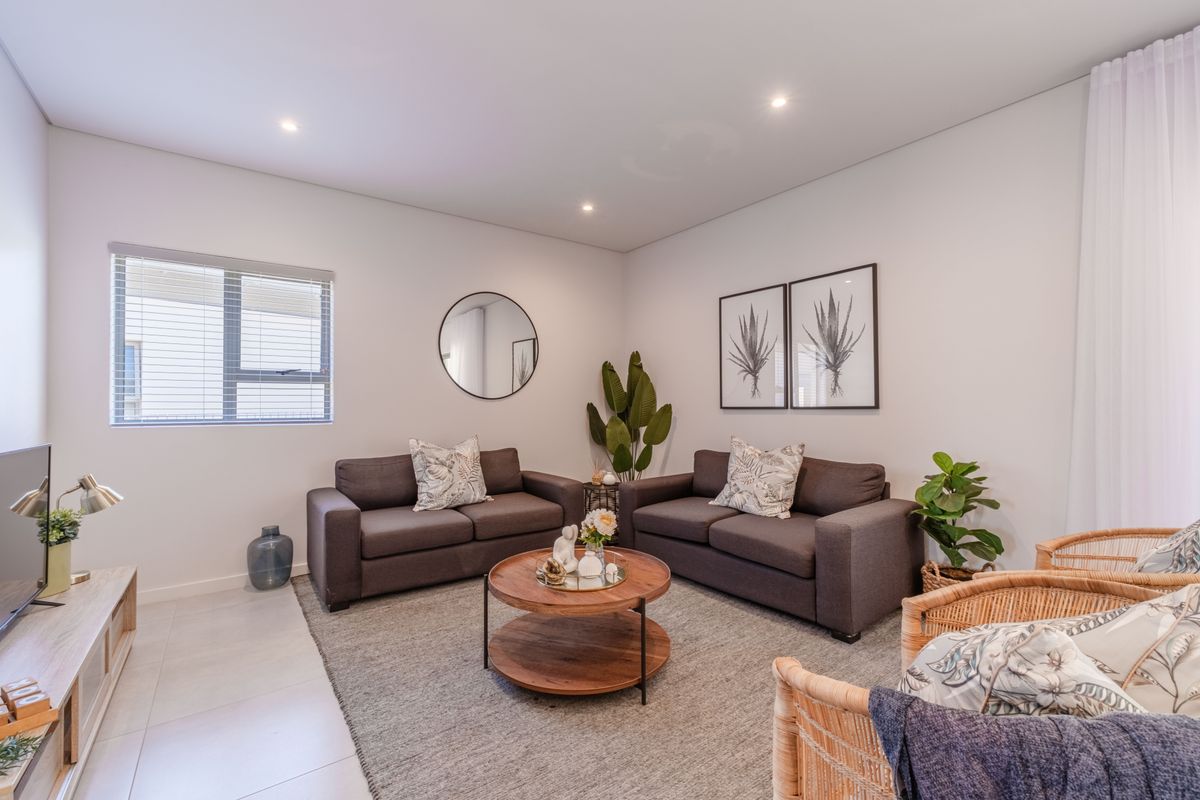 A bright and modern living room featuring neutral tiled flooring, recessed downlighting, and large windows with fitted blinds.