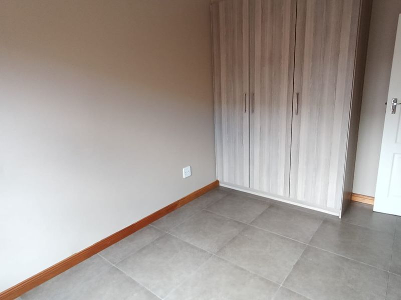 A clean, empty bedroom featuring neutral wall tones, modern grey floor tiles, and a functional built-in wardrobe with wood-grain finish.