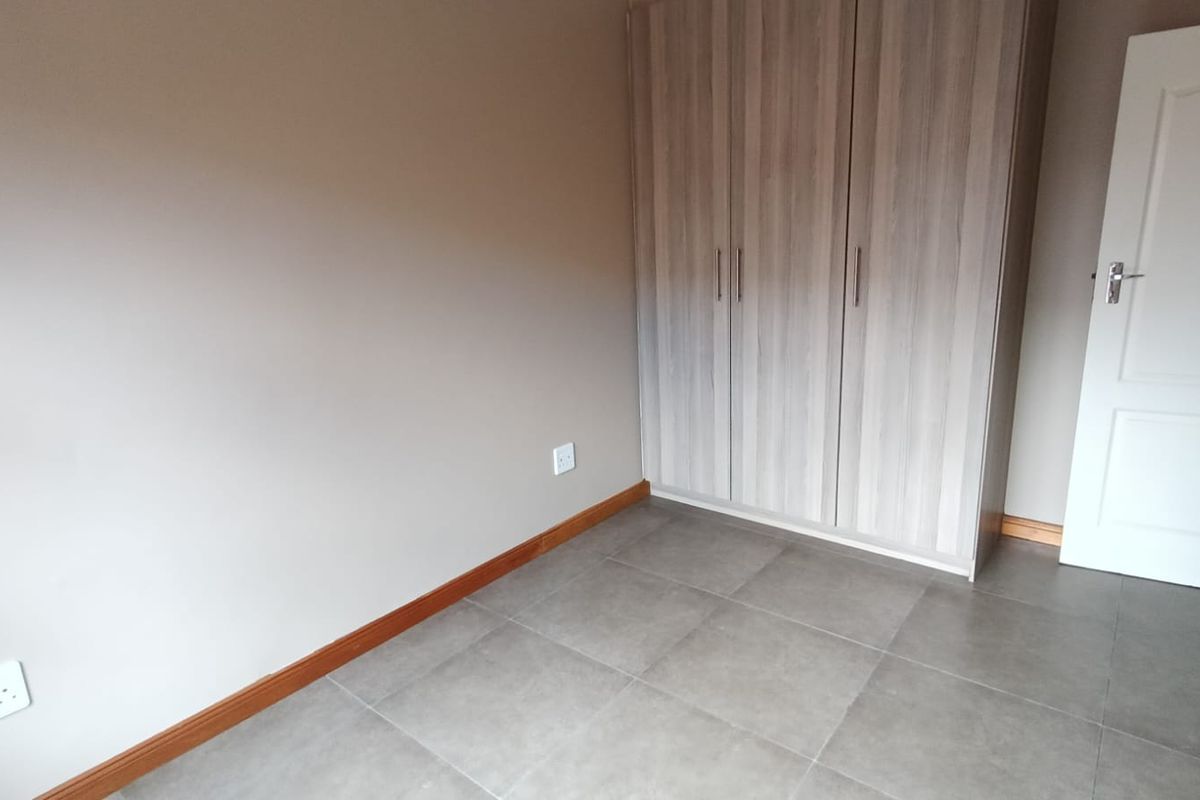 A clean, empty bedroom featuring neutral wall tones, modern grey floor tiles, and a functional built-in wardrobe with wood-grain finish.