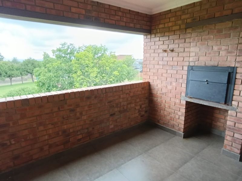 A covered balcony featuring exposed brick walls, tiled flooring, and a built-in braai, offering an open view of the surrounding greenery.