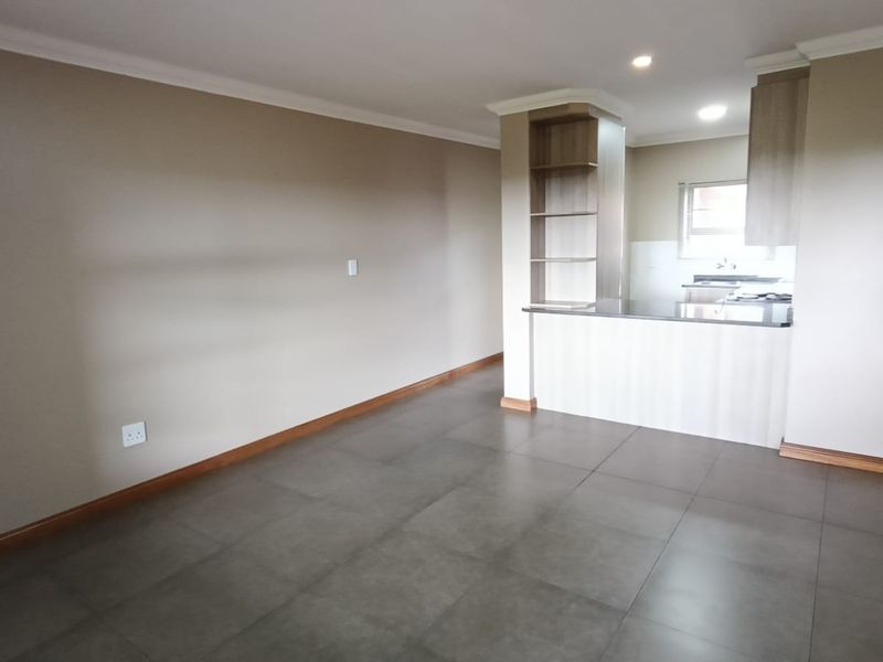 An open-plan living and kitchen area featuring large format floor tiles, neutral wall tones, and a functional breakfast bar with integrated shelving.
