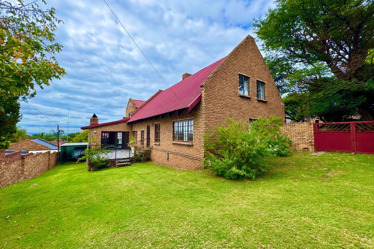 A rear view of a traditional brick house featuring a pitched red metal roof, a paved patio area, and a spacious surrounding lawn.