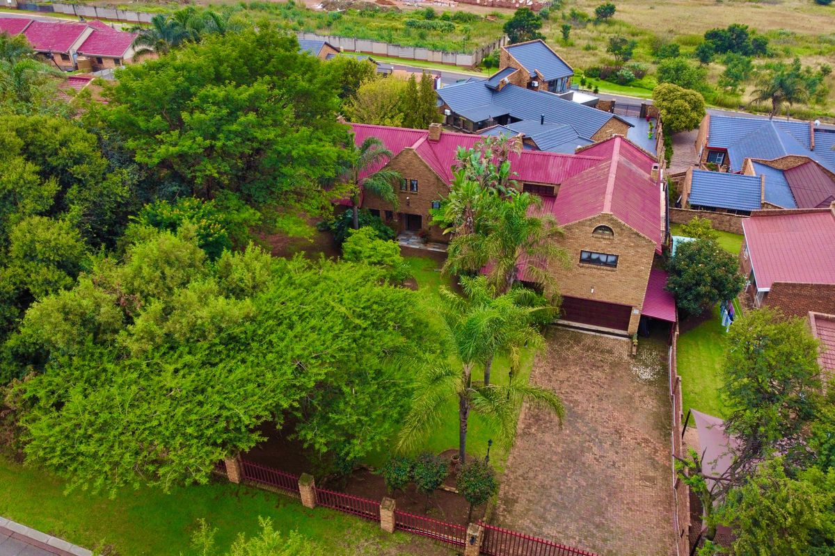 Aerial view of a brick house with a red metal roof, featuring a paved driveway, established garden, and perimeter fencing in a residential estate.