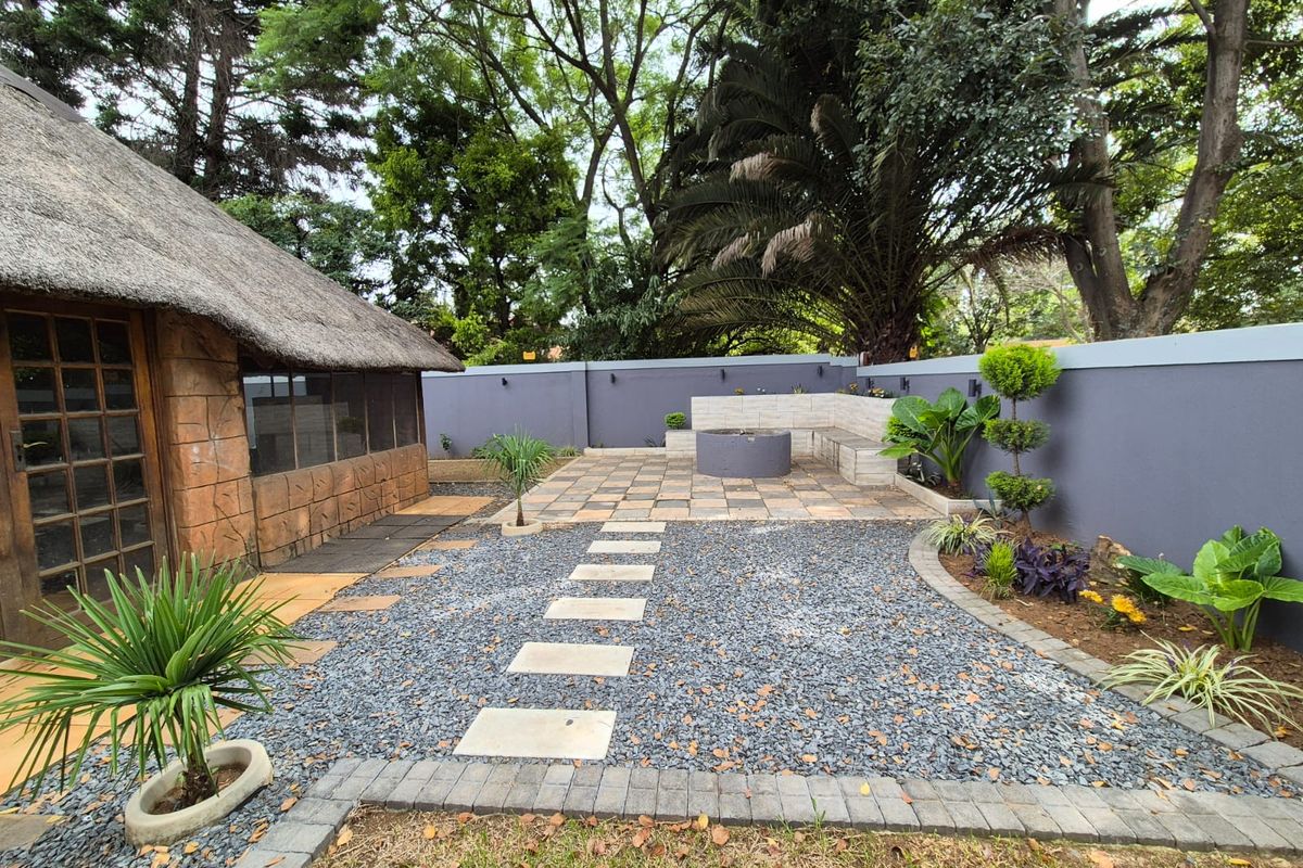 A landscaped rear garden area featuring a thatched structure, paved patio with a built-in fire pit, stone pathways, and perimeter boundary walls.