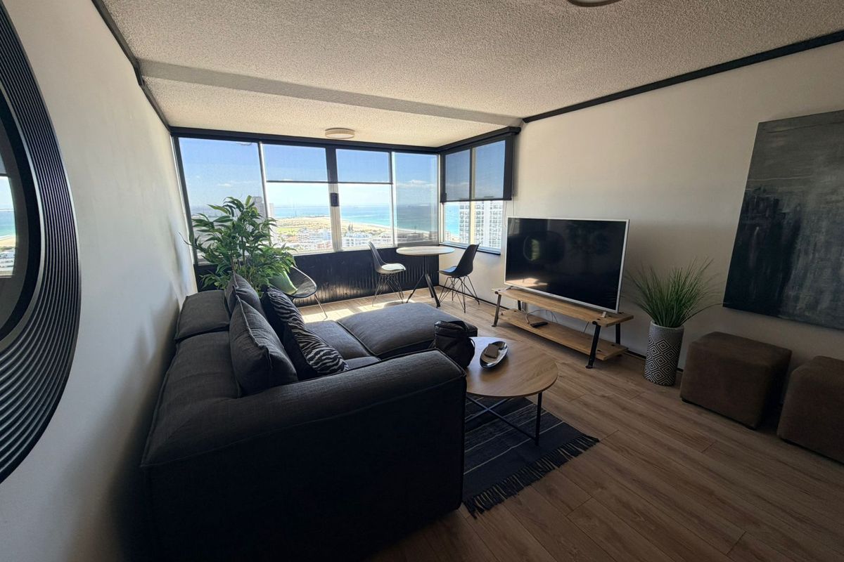 A bright living room featuring large floor-to-ceiling windows with expansive coastal views, wood-look flooring, and a functional open layout.