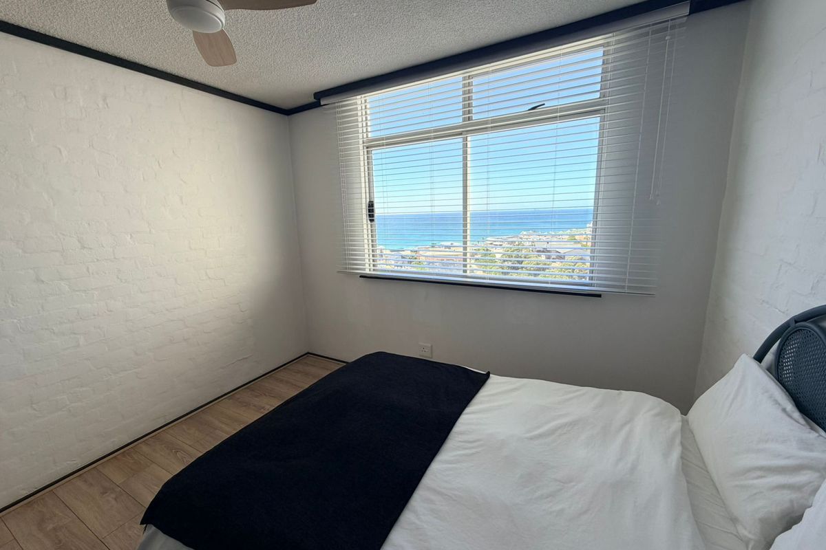 A bedroom featuring light wood-look flooring, white painted brick walls, and a ceiling fan, offering a clear view of the ocean through the window.