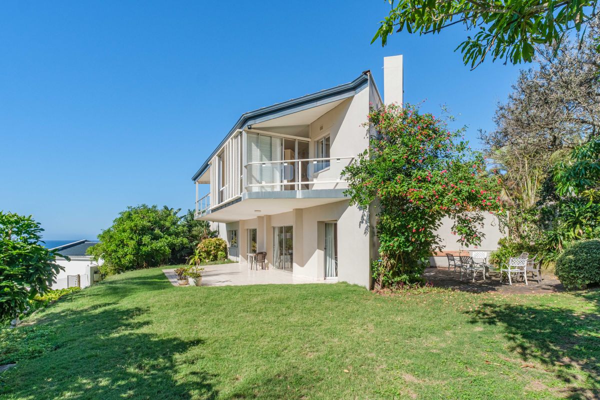 Rear view of a multi-level house featuring a balcony, paved patio area, and established garden with ocean views in the distance.