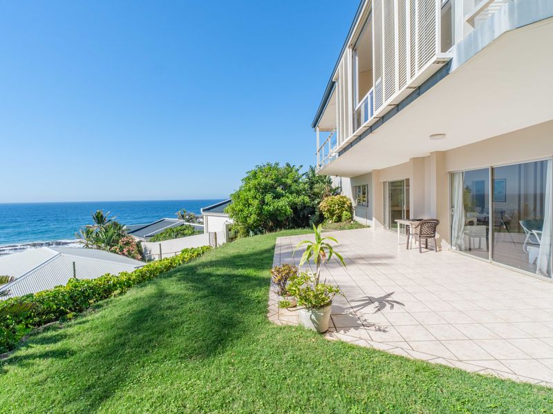 A spacious covered patio overlooking a manicured lawn with expansive ocean views, featuring large sliding glass doors providing seamless indoor-outdoor flow.
