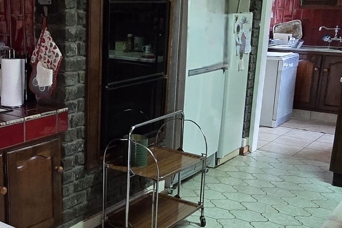 A dated kitchen featuring exposed brick walls, built-in oven, and tiled flooring, with an adjacent scullery area visible through the doorway.