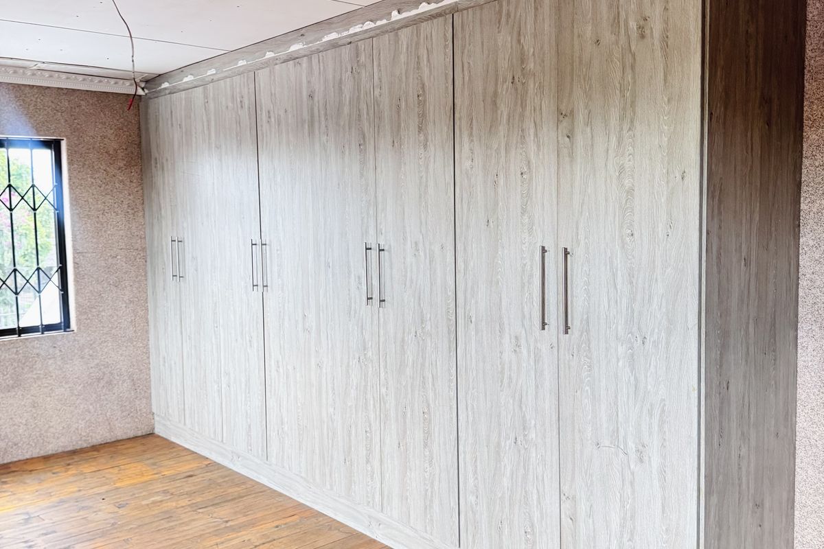 A bedroom featuring large built-in wooden wardrobes, wooden flooring, and a security-barred window, currently undergoing renovation with unfinished ceiling and trim work.