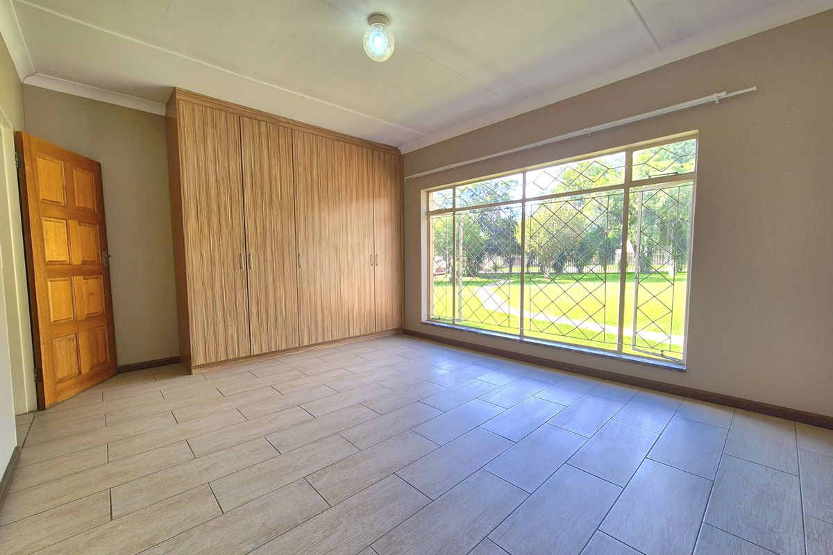 A bright, empty bedroom featuring wood-look floor tiles, built-in wooden cupboards, and a large window with security bars overlooking the garden.