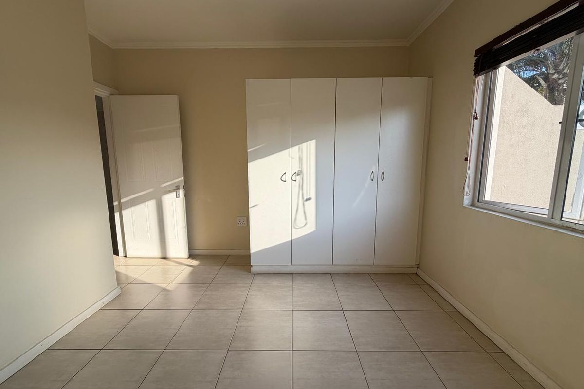 A bright, empty bedroom featuring neutral tiled flooring, built-in white cabinetry, and a window with fitted blinds.