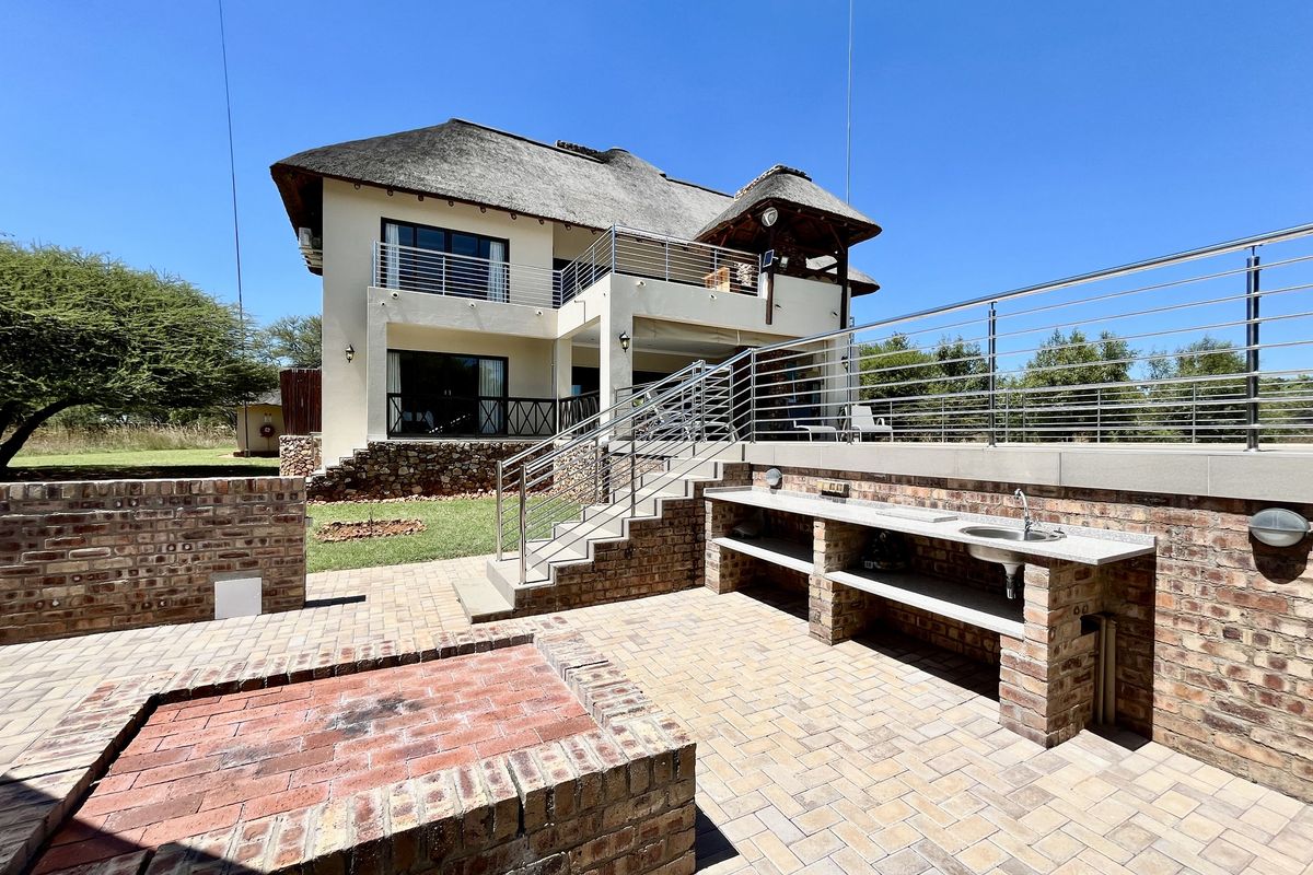 Spacious outdoor entertainment area featuring a built-in brick braai, stone-topped prep station with sink, and a raised patio overlooking the rear garden.