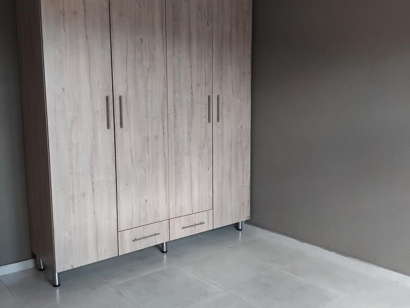 A clean, empty bedroom featuring light wood-finish built-in wardrobes, neutral grey walls, and modern tiled flooring.